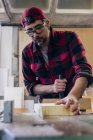 Carpintero en gafas de seguridad cortando pieza de madera en taller - foto de stock