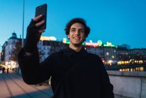 Улыбающийся мужчина в теплой одежде стоит на вечерней улице и делает селфи со смартфоном . — стоковое фото