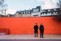 Двое мужчин в теплой одежде стоят у оранжевой стены на улице и смотрят в сторону — стоковое фото