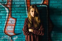 Красивая молодая женщина со скрещенными руками позирует на кирпичной стене с граффити и смотрит в камеру — стоковое фото