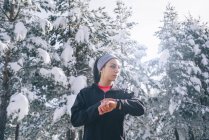 Sportive woman holding hand with sports watch at winter forest and looking aside — Stock Photo