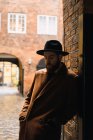 Stylish  man in coat and hat leaning on archway wall and looking down — Stock Photo