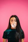 Portrait of woman looking at camera and blowing gum bubble on pink background. — Stock Photo