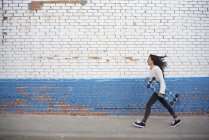 Side view of brunette girl walking with longboard along street wall — Stock Photo
