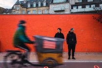 Rickshaw-puller ridding along two men in warm clothes standing by orange wall on street scene. — Stock Photo