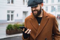 Усміхнений бородатий чоловік у кепці, використовуючи смартфон на вулиці — стокове фото