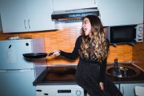 Жінка сміється, сидить на кухонному столі і витягує руку зі сковородою — стокове фото