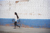 Seitenansicht eines brünetten Mädchens, das mit Longboard auf der Straße läuft — Stockfoto