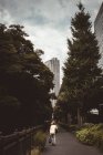 Visão traseira da mulher andando no beco com bosques altos no fundo da cidade moderna . — Fotografia de Stock
