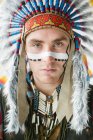Молодой человек с чертой на лице позирует в традиционном индейском костюме и смотрит в камеру — стоковое фото