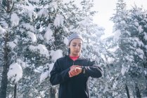 Портрет здоровой женщины, смотрящей на спортивные часы в зимнем лесу — стоковое фото
