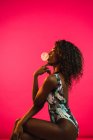 Side view of pretty black woman in bodysuit posing and chewing gum on pink background. — Stock Photo