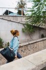 Vista lateral de la chica en chaqueta de mezclilla caminando con bolsa de viaje en el pasaje urbano - foto de stock