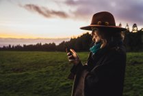 Femme souriante dans le chapeau navigation smartphone aux champs froids verts au crépuscule — Photo de stock