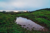 Small pond reflecting cloudy dark sky in green fields in highlands. — Stock Photo