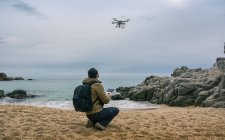 Вид сзади человека с рюкзаком, испытывающего дрон в воздухе на пляже — стоковое фото