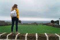 Visão traseira da mulher com mochila em pé na cerca no campo verde no dia nublado . — Fotografia de Stock