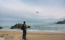 Вид ззаду чоловіка, що стоїть на пляжі і тестує безпілотник в повітрі — стокове фото