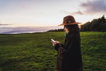 Vue latérale de la femme dans le chapeau navigation smartphone dans les champs verts — Photo de stock