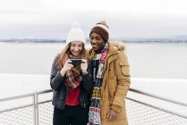 Cheerful couple standing at fence and looking in smartphone against ocean. — Stock Photo
