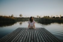Вид сзади женщины, сидящей на деревянном пирсе у озера — стоковое фото