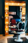Empty chair placed at mirror with illumination in barbershop. — Stock Photo