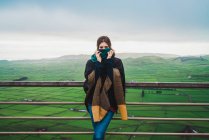 Mulher escondendo rosto com cachecol e inclinando-se na cerca rural no fundo da vista panorâmica incrível de campos enevoados — Fotografia de Stock