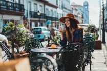 Улыбающаяся молодая женщина в шляпе сидит с чашкой на террасе кафе . — стоковое фото