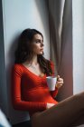 Mulher pensativa em vestido vermelho bebendo café em casa — Fotografia de Stock