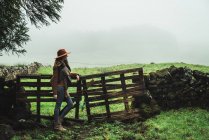 Vista lateral de la mujer en chaqueta y sombrero apoyado en la cerca vieja con campos brumosos en el fondo . - foto de stock