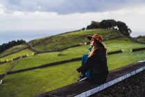 Joven mujer soñadora sentada en la cerca en el fondo del campo verde en la colina . - foto de stock