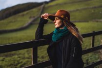 Mulher sonhadora de chapéu em pé na cerca e olhando para longe no campo verde . — Fotografia de Stock