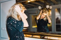 Young blonde woman looking at camera and holding head while reflecting in mirror. — Stock Photo