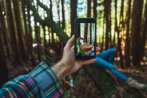 Hand mit Smartphone fotografiert hübsche Frau beim Entspannen auf Baumstamm im sonnigen Wald. — Stockfoto
