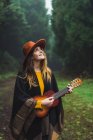 Jovem mulher sorridente de chapéu tocando guitarra pequena e olhando para cima em bosques nebulosos verdes . — Fotografia de Stock