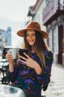 Усміхнена жінка в капелюсі, сидячи за столом тераси кафе з чашкою і смартфоном в руках — стокове фото