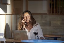 Женщина разговаривает по телефону, сидя в кафе с вином и ноутбуком — стоковое фото