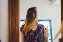 Woman standing at mirror with sunglasses over hair and gesturing V-sign — Stock Photo
