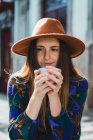 Mujer sonriente en sombrero posando con taza en la cafetería exterior - foto de stock