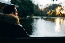 Back view of man in warm jacket sitting and looking at lake in sunset. — Stock Photo