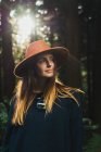 Young pretty woman in hat standing in sunny forest and looking away. — Stock Photo