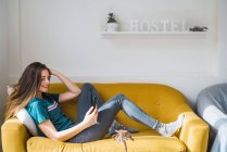 Весела дівчина з телефоном, що розслабляється на дивані на фоні стіни з вимощенням хостелу на полиці — стокове фото