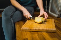 Plan de la culture de la femme assise sur le comptoir et couper morceau d'ananas avec couteau tranchant . — Photo de stock
