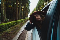 Жінка в капелюсі висить з машини на лісовій дорозі — стокове фото