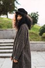 Вид збоку досить кучерявої жінки в стильному пальто позує в міському парку . — стокове фото