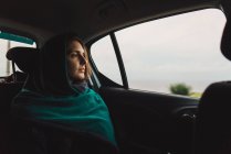 Femme en écharpe chevauchant sur le siège arrière dans la voiture et regardant de rêve dans la fenêtre . — Photo de stock