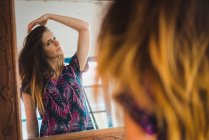 Junge Frau steht vor Spiegel und schaut auf Spiegelung, während sie die Haare verstellt. — Stockfoto