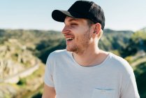 Smiling young man in cap standing in mountains in sunny day. — Stock Photo