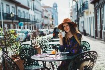 Усміхнена жінка з чашкою, що сидить за столом тераси кафе — стокове фото