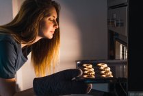 Vista lateral da menina verificando o progresso de fazer biscoitos na panela da folha no forno . — Fotografia de Stock